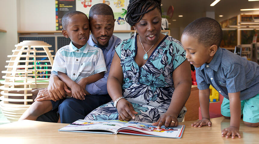 A family comprised of a mother, father and two young boys read a book together in the Children's section of the library.