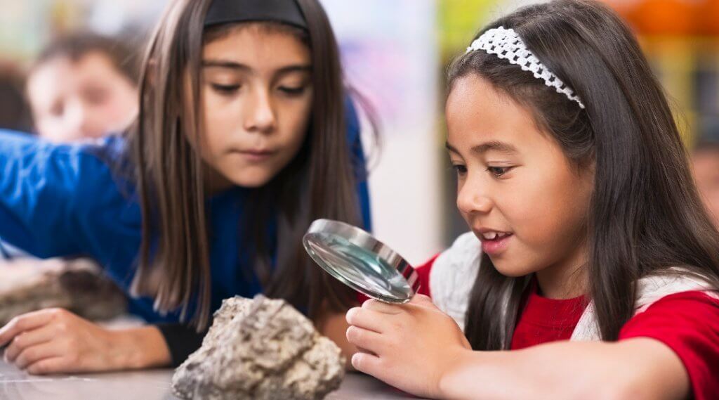 Two children use a magnifying glass to inspect a rock.