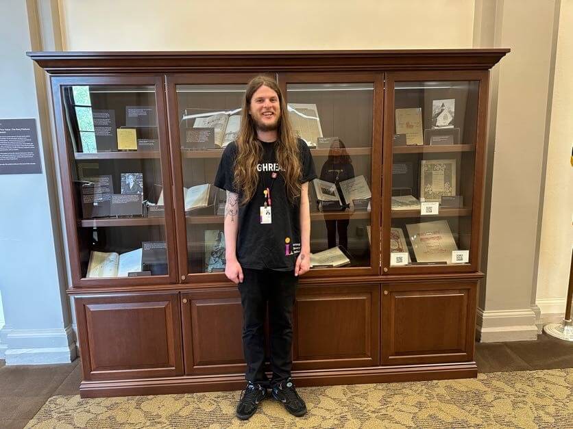 Smiling young white man with long hair and beard, standing in front of a glass and wood display case