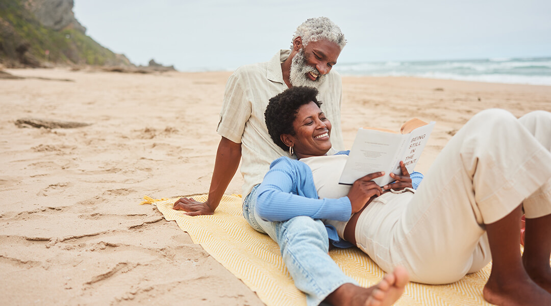 Smiling mature adult reading a book while lying on their partner's lap during a day on a sandy beach.