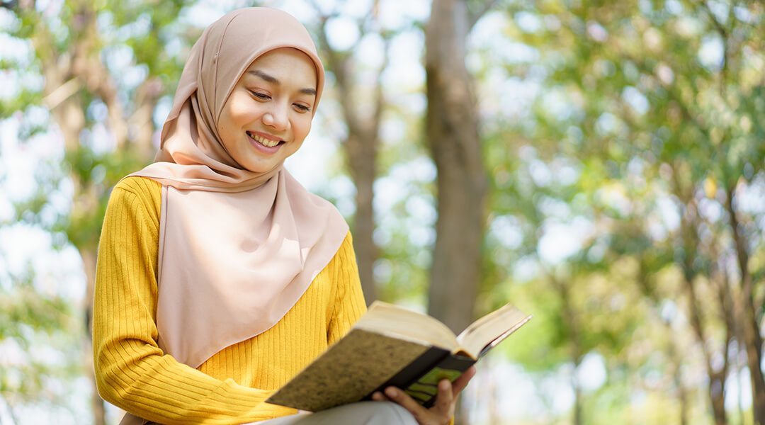 Adult wearing a hijab reading a book outside.