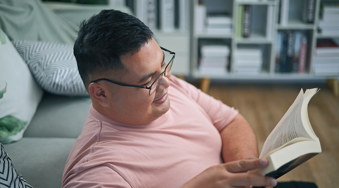 An adult wearing glasses sits on the floor, reading a book.