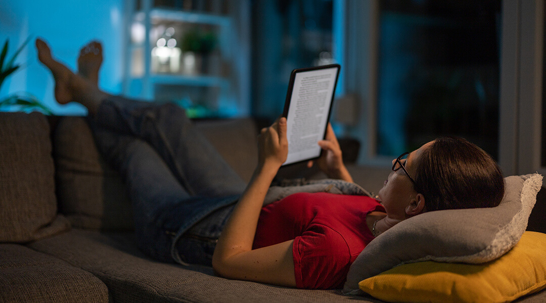 Adult lying on sofa in living room at night, and reading a book on digital tablet in peace.