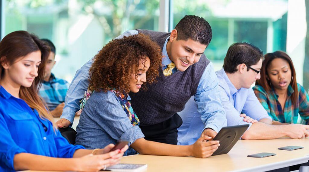 A librarian helps a patron use a tablet, in a class with other adult students and their digital devices.