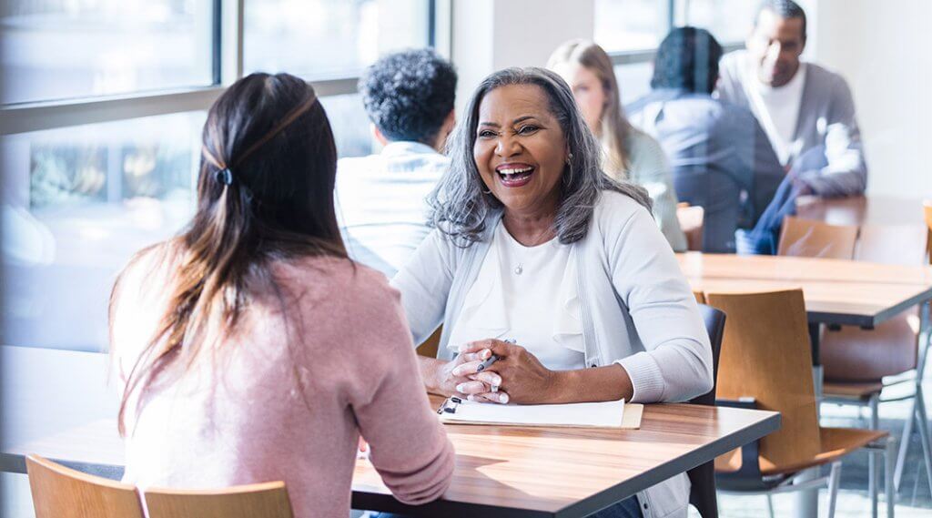 Two adults share a table in a community room, one smiling and poised with a clipboard as they talk to the other person.