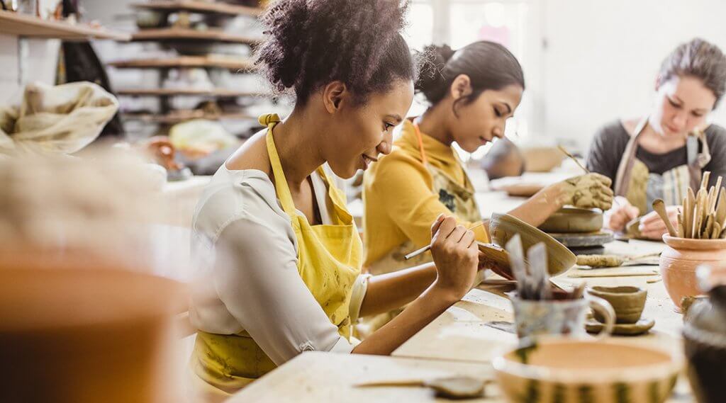 Three young adults mold and paint pottery in an art classroom.