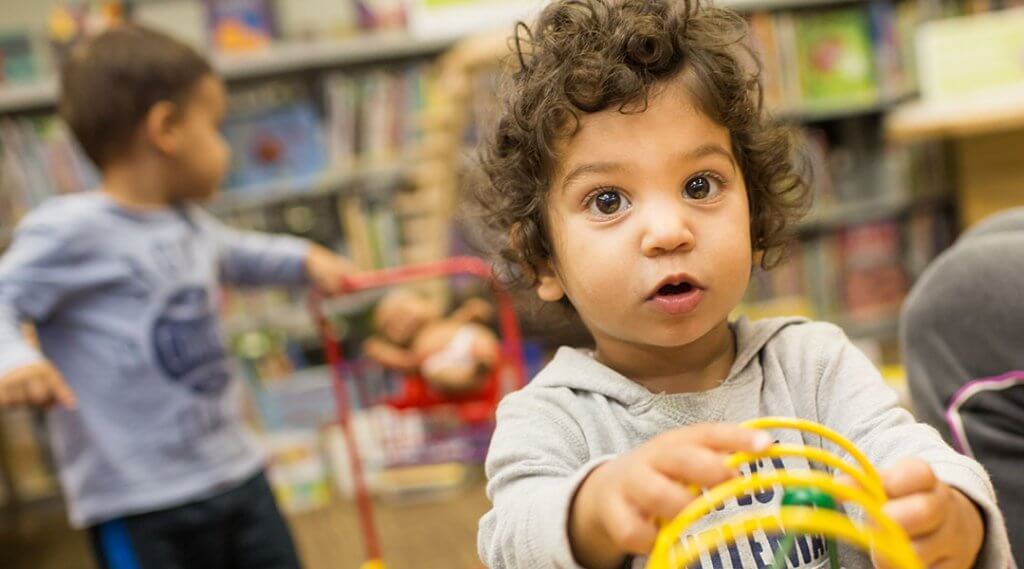 Close-up of a toddler playing with a bead table, with another young child playing in the background.