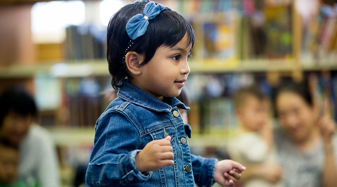 Medium shot of a toddler standing up during storytime, with other childrens and caregivers out of focus in the background.