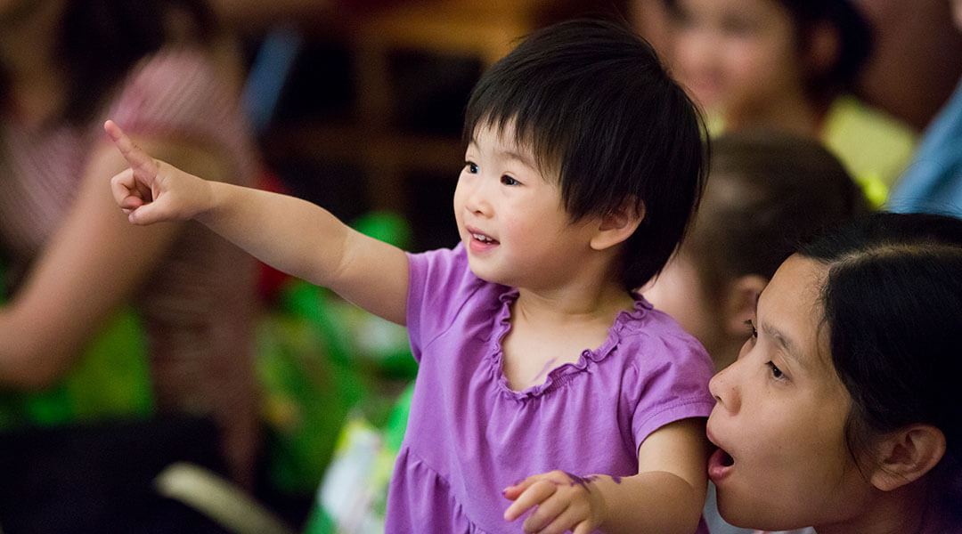 A young child excitedly points forward, supported by their caregiver, with other storytime audience members in the background.