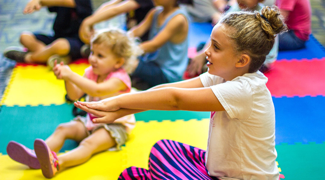 Young children of varying age stretch while sitting on a colorful mat during storytime.