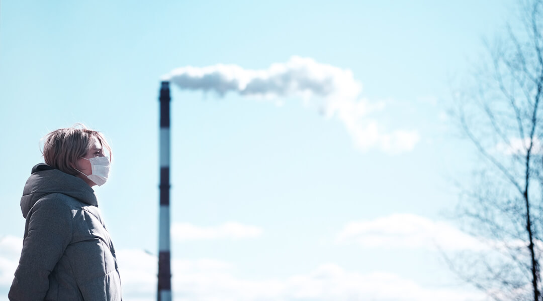 A person wearing a coat and face mask stands in front of a blue sky with a single smoke stack producing white smoke in the air.