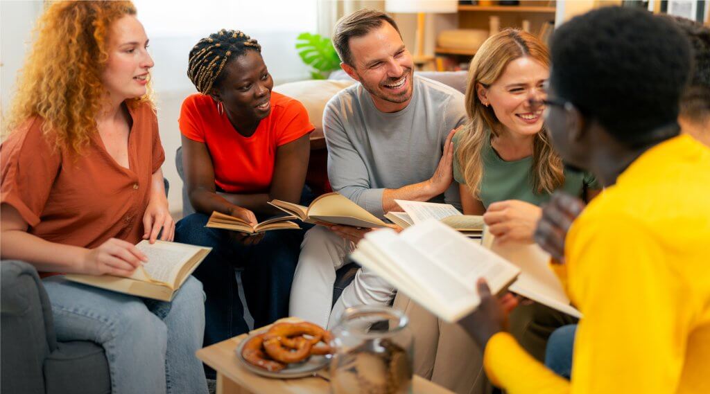 A diverse group of adults sitting together around a small table with refreshments, smiling and holding open books.