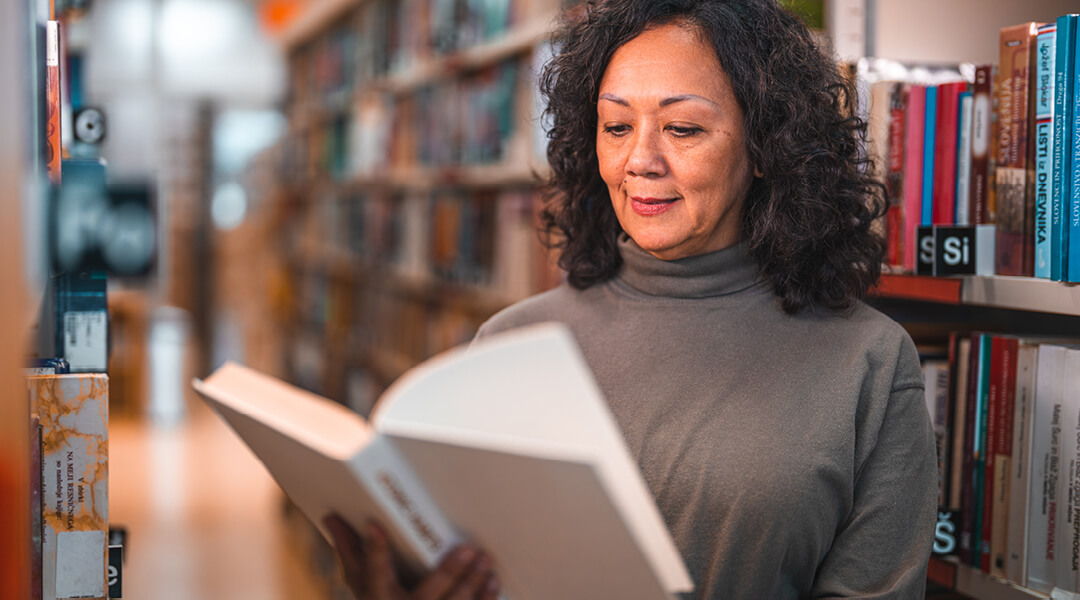 Woman standing in front of library bookshelves reading a book.