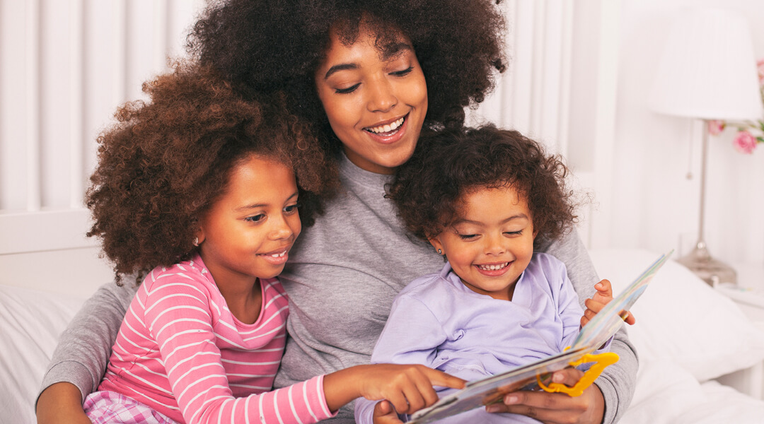 Adult and 2 young children, all with the same hair style, cuddling and smiling, reading a book together,