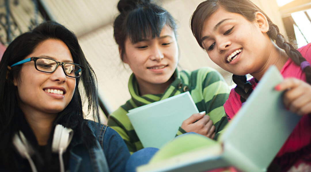 Three teenage girls smiling and looking at a book together.