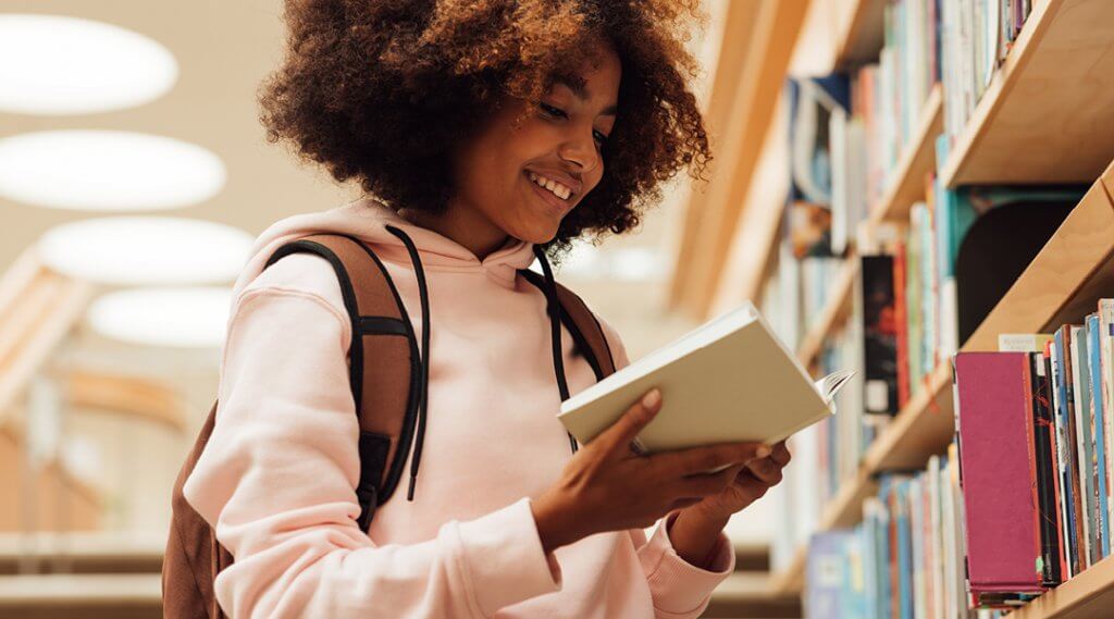 Teenager smiling while reading a book in front of a library bookshelf.