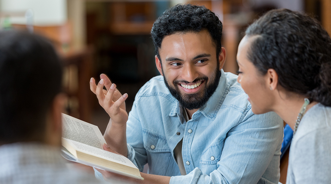 A smiling young adult discusses a book with a friend in the library.