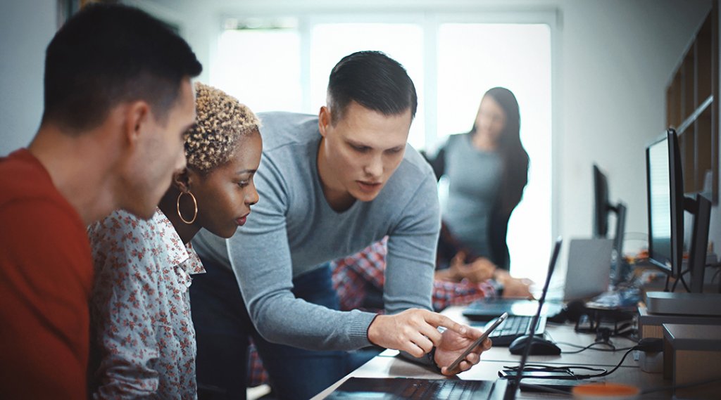 A person points to their phone screen to show two coworkers an issue in a computer lab.