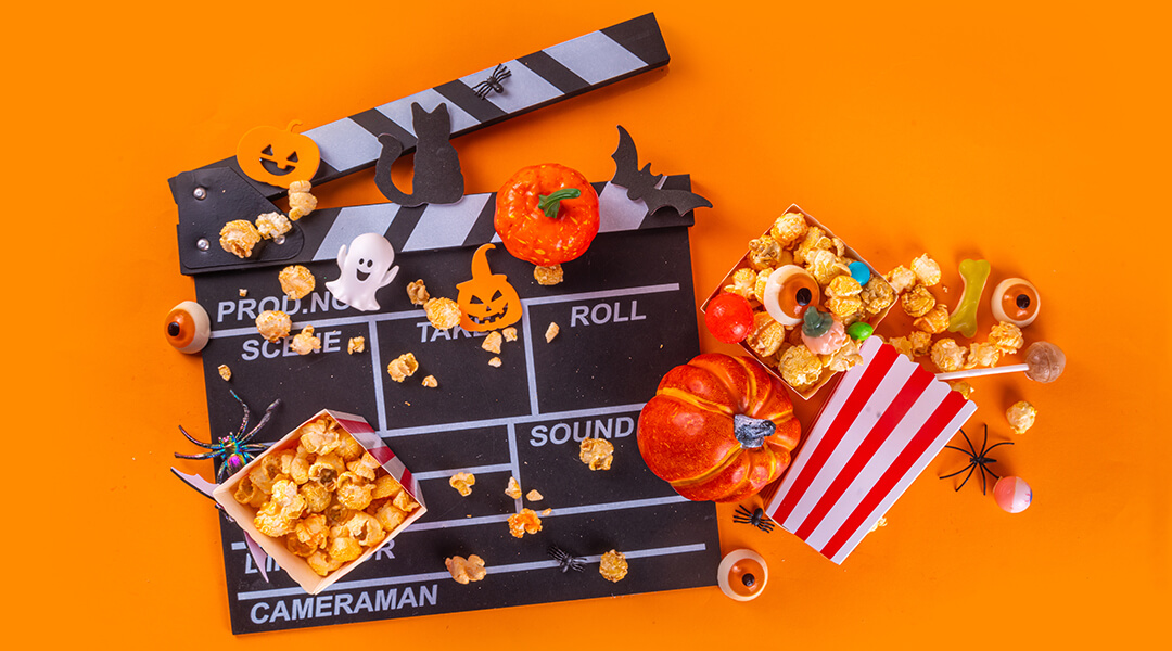 Trick-or-treating sweets bucket, popcorn, movie clapperboard and Halloween decorations on orange background.