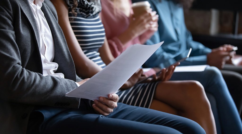 Close up unrecognizable young multiracial people sitting in line on sofa with paper documents waiting for job interview.