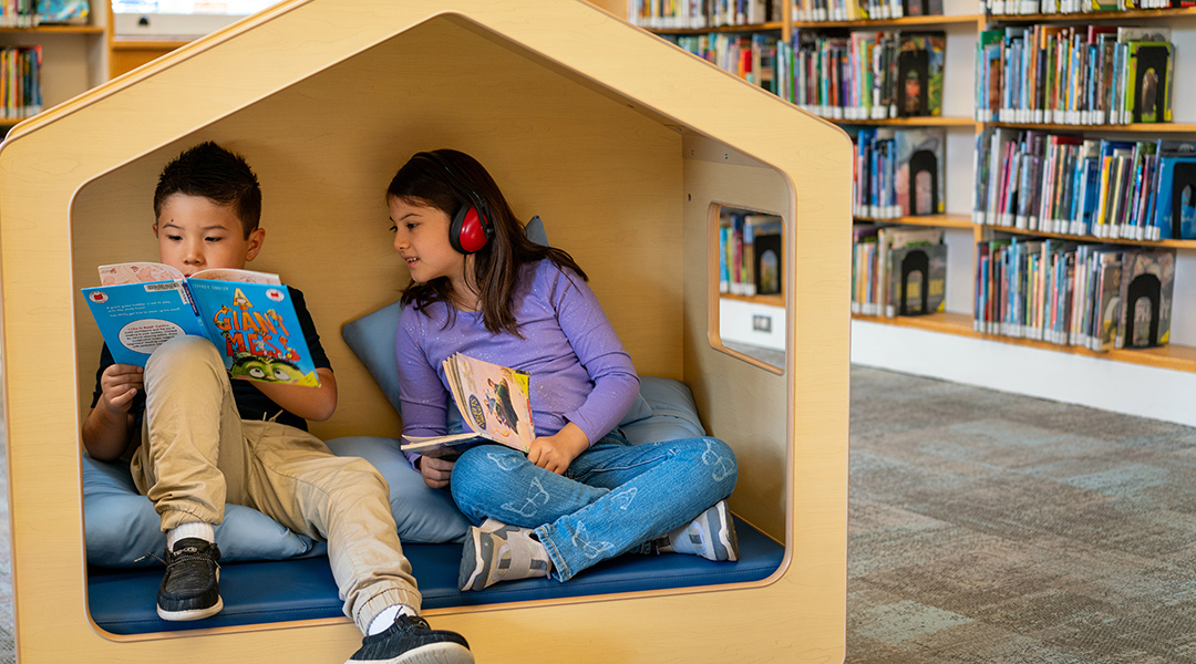 Two siblings, about 5 and 8, read together in a cubby in the children's section of the library. The older child is wearing sensory headphones.