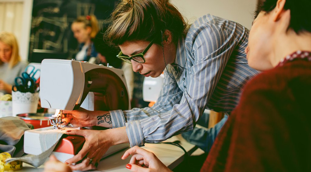 An instructor helps a student on a sewing project, adjusting the foot of the sewing machine.