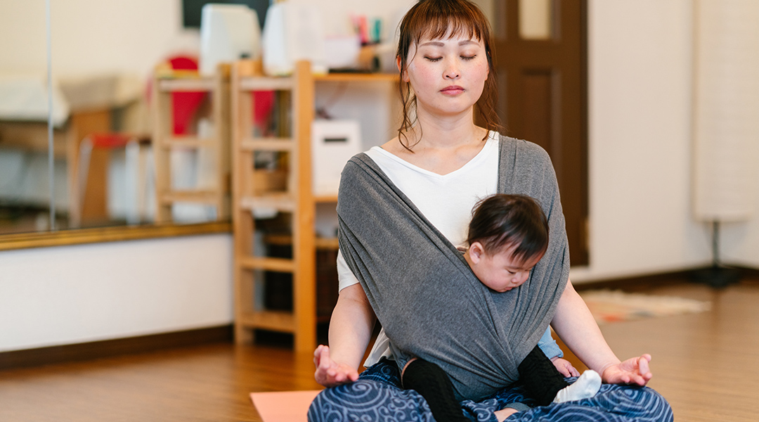 A mother is practicing yoga and meditating while carrying her baby in a baby fabric sling.