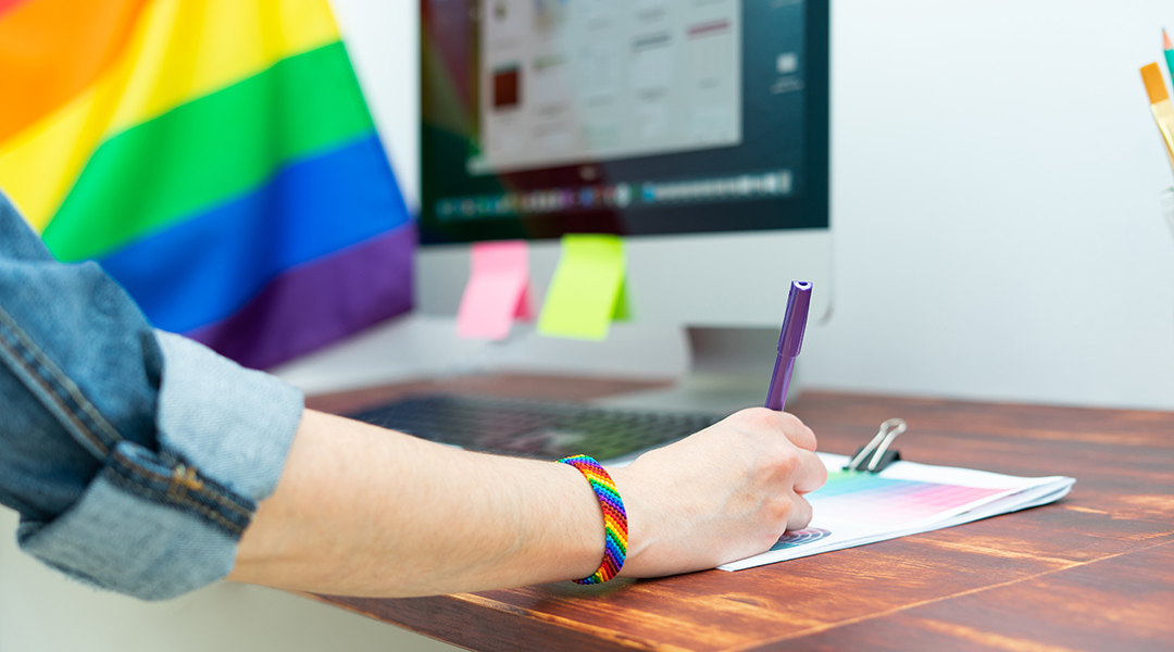 Close-up of a person's hand taking notes near a computer; there is a pride flag in the background and the person is wearing a rainbow friendship bracelet.