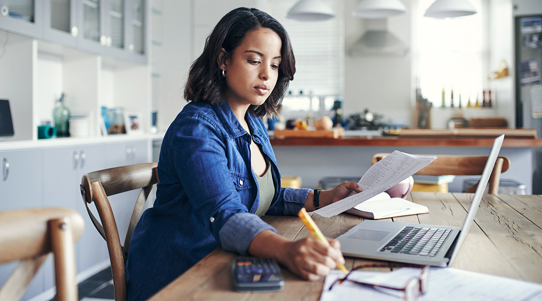 A young adult using a laptop and reviewing documents with a calculator at home.