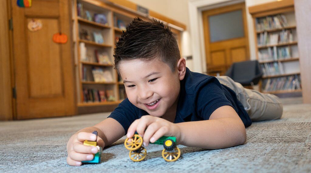 A young child lays on their stomach in the library, playing with a toy wagon.