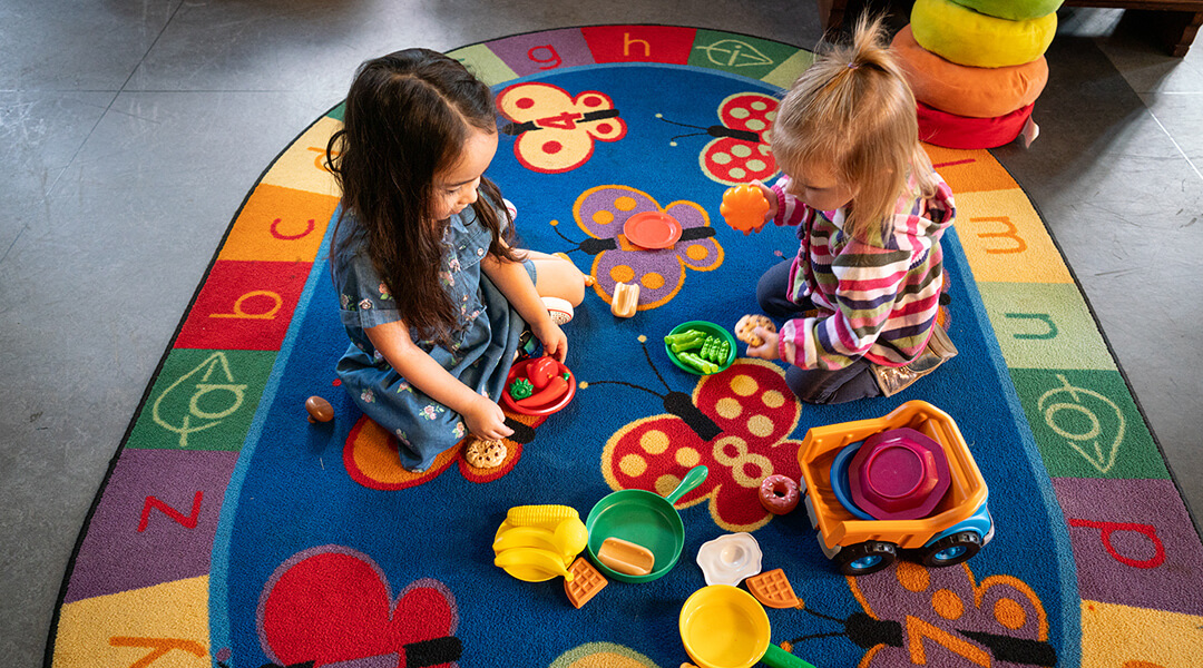 Two toddlers play with toy food, sitting in the center of a colorful rug.