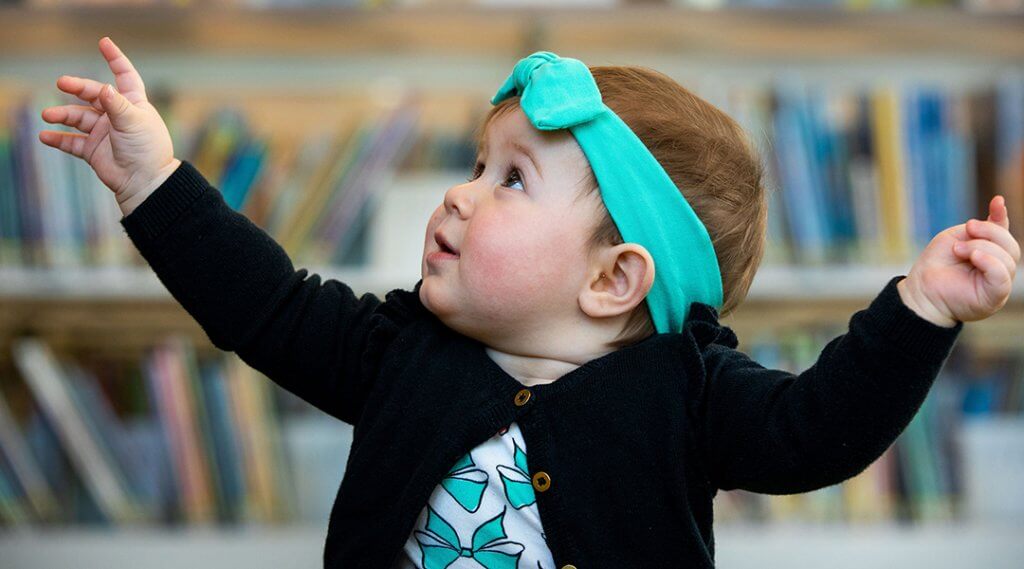 Close-up of a baby looking up and out of frame with both hands raised.