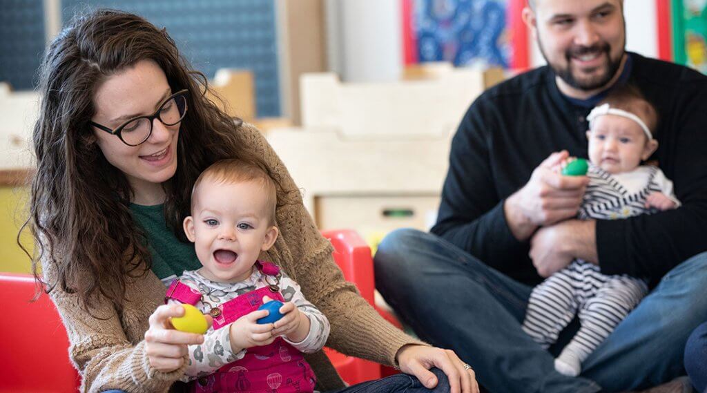 Two babies sit with their caregivers playing with shaker eggs during storytime.