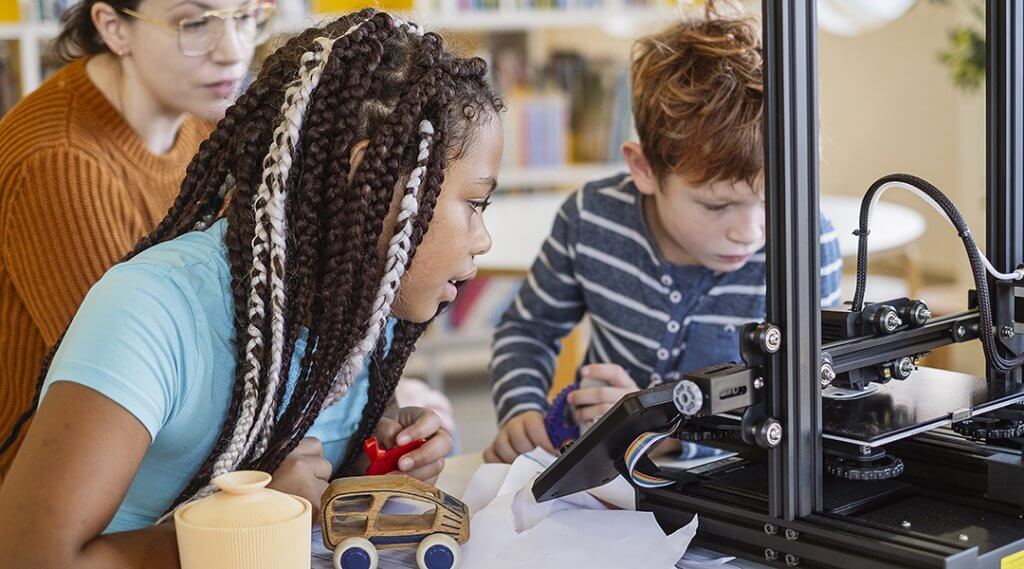 A librarian helps two children use a 3d printer in the library.