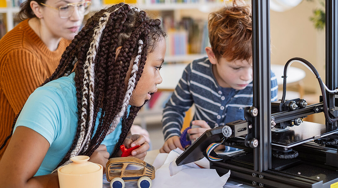 A librarian helps two children use a 3d printer in the library.