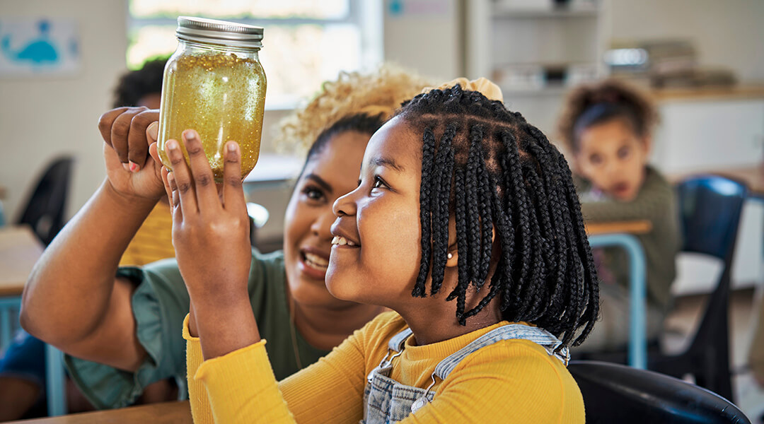 An elementary school age child and teacher admire the experiment of a glittery substance in a mason jar.