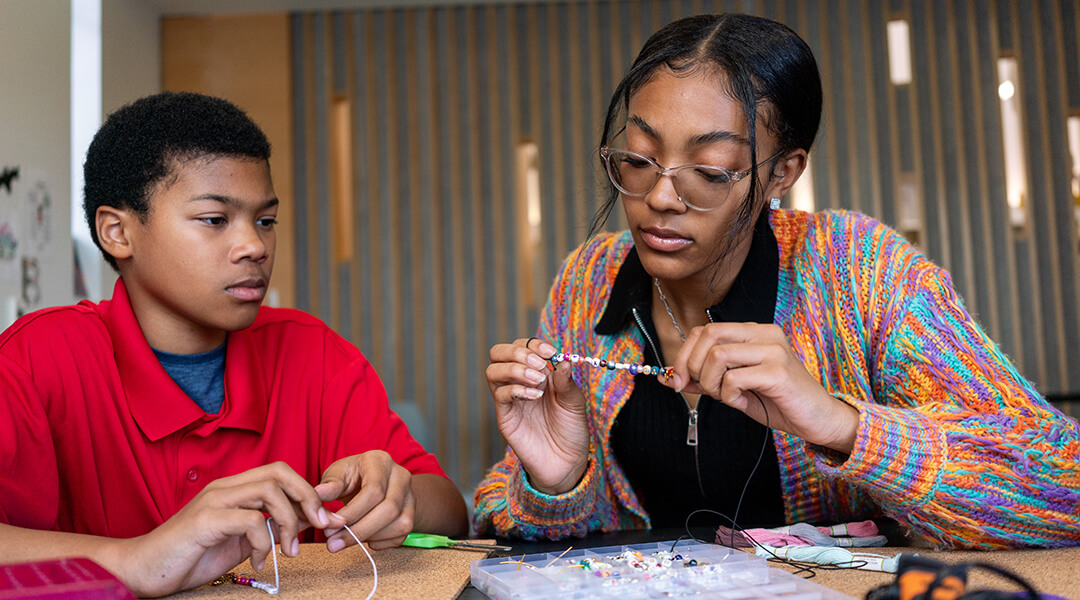 Two teenagers sit at a table beading friendship bracelets.