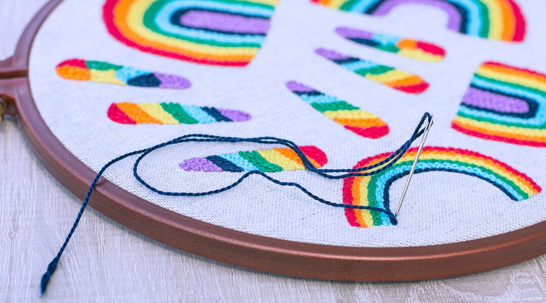 Closeup of needle with blue thread in grey fabric with embroidered rainbows of different shapes and sizes.