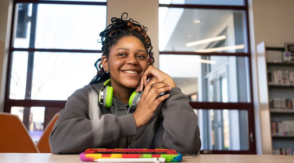 A smiling teen sitting at a table in the TeenSpace, with a tablet in front of them.