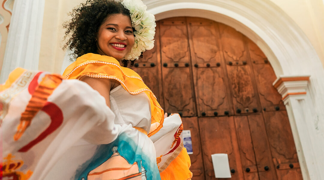 A woman in a colorful dress and floral hairpin smiles in front of an arched wooden door.