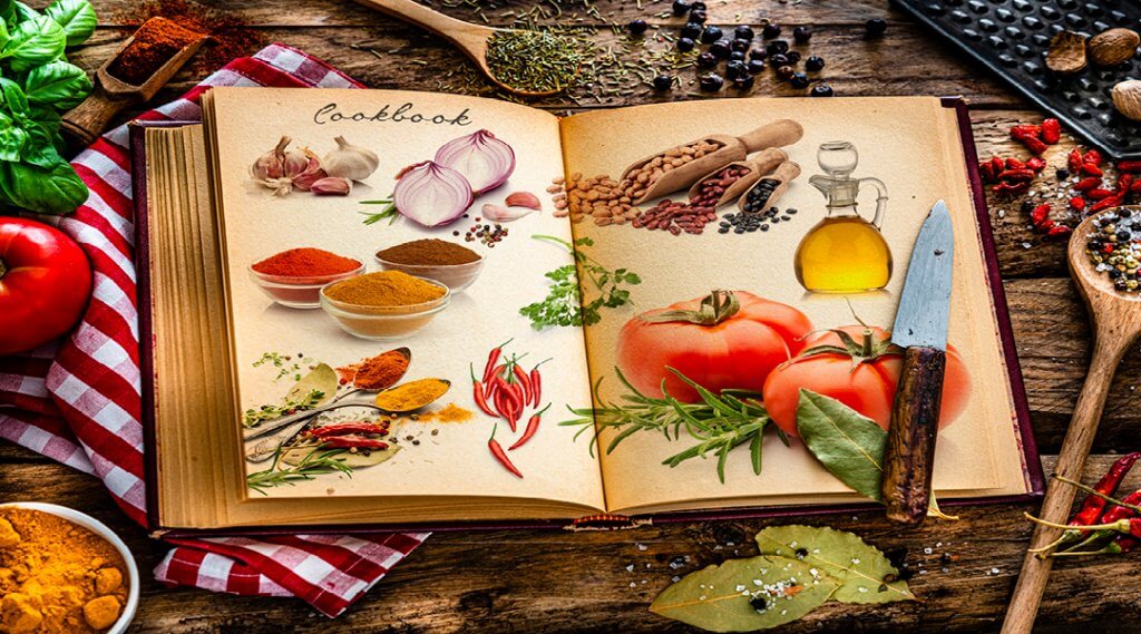 A cookbook open to pages of illustrated ingredients on a table surrounded by cooking supplies.