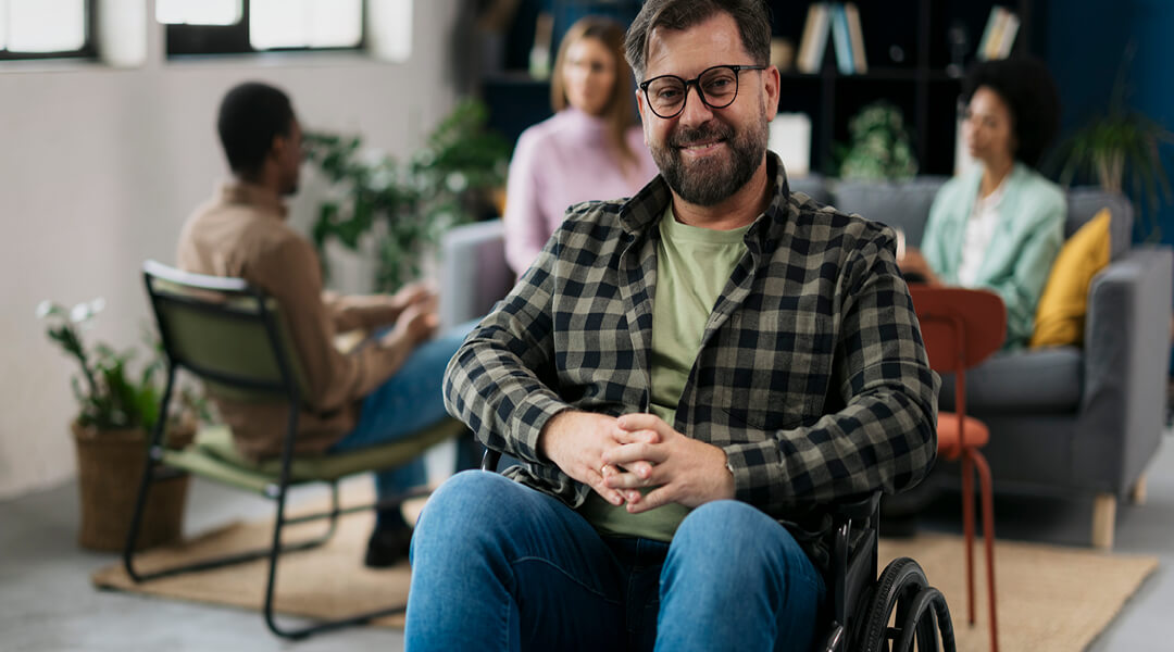 An adult using a wheelchair smiles with three other people out of focus in the background.