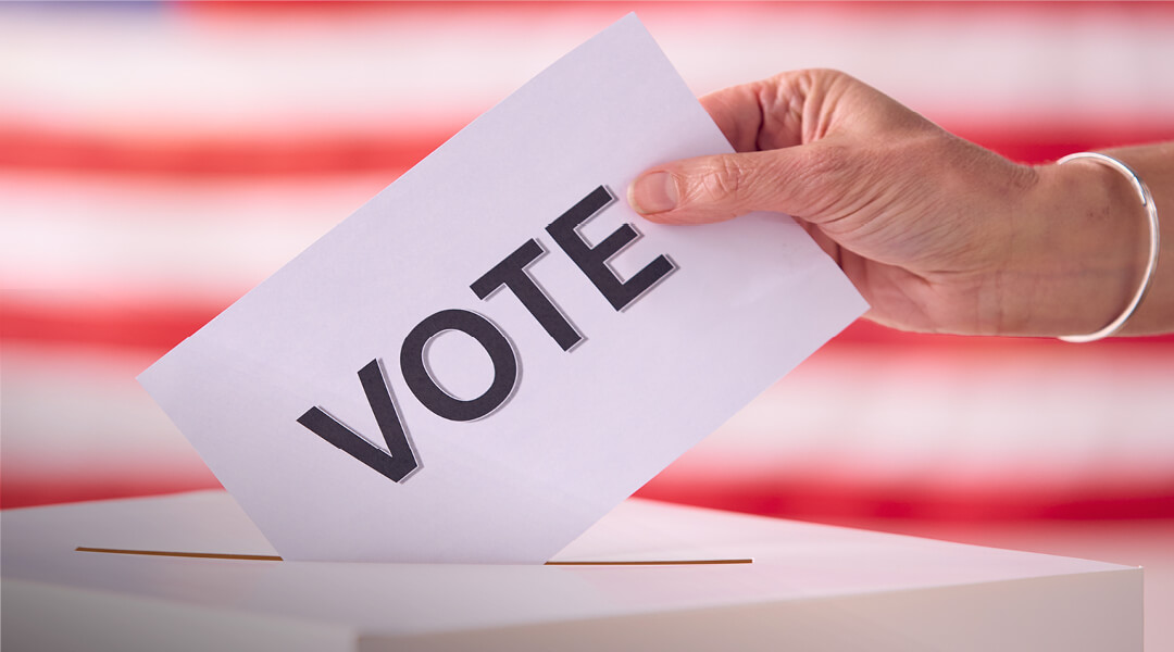 Close-up of a hand putting a piece of paper that reads, 'vote' in a simple ballot box.