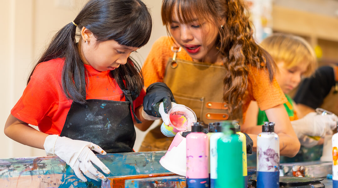 A teenager helps a child, about ten, pour paint for a craft project.