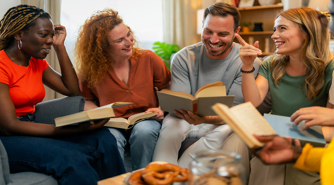 Four smiling adults laugh together on a couch while discussing the books they're holding.