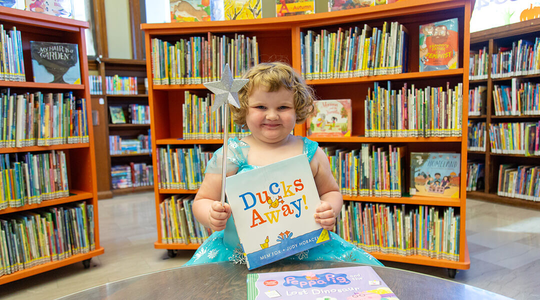 Smiling little girl with a silver wand holding up a book in the Children's Department