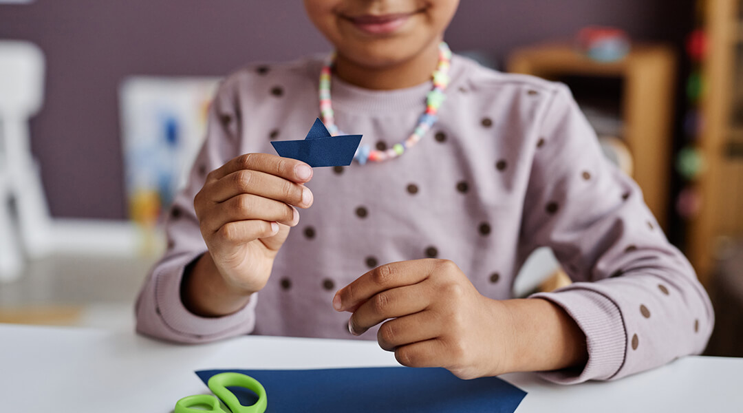 Close-up of cute little learner holding tiny origami ship.