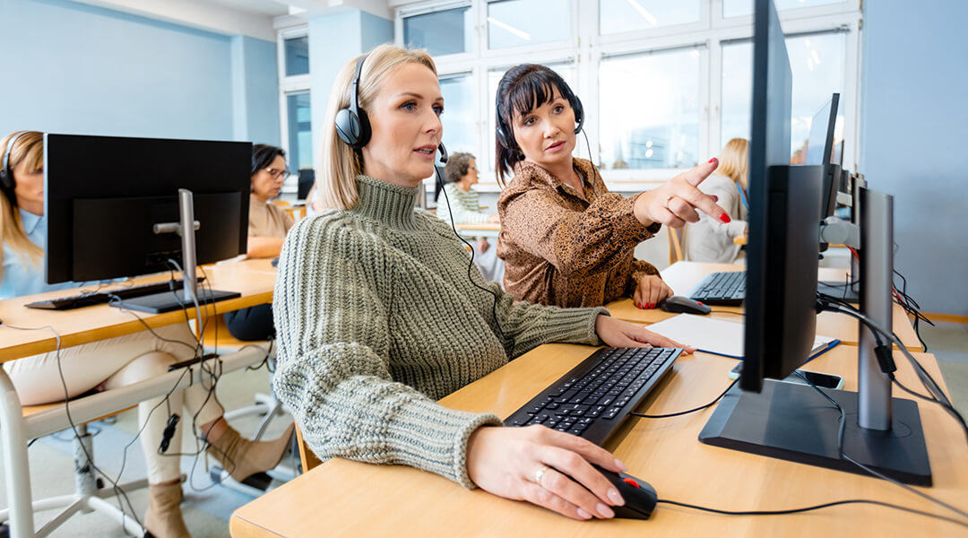 A group of adults at computers with headphones on. The person in the center is being assisted by an instructor.