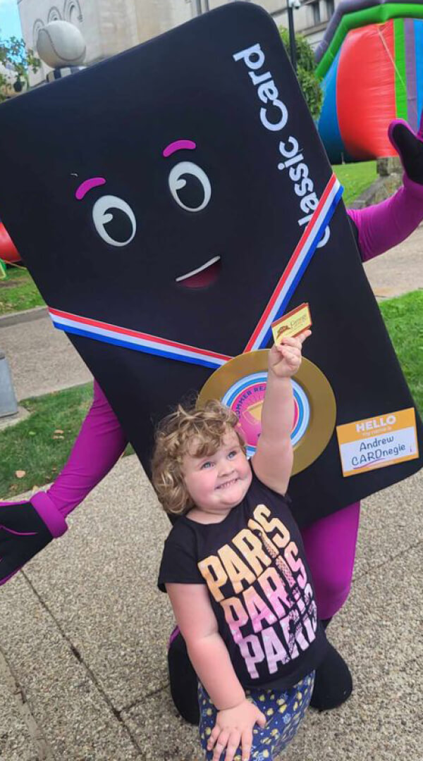 A toddler, Pim, holds hands with CLP's mascot Andrew CardNegie at a summer reading event.