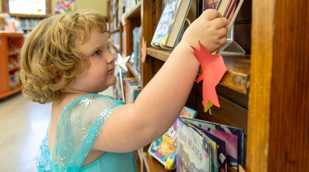 A toddler in a blue princess dress explores a shelf of books on display in the library's children's section.